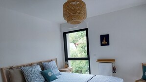 A bedroom with a bed, a window, a wall-mounted heater, and a wooden nightstand. - Luxury Lakeside T3: Modern, Bright, Near Beach, WiFi, Parking (Soorts-Hossegor)