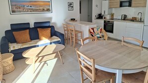 A modern living room with a blue sofa, a round wooden table, and a kitchen area with white cabinets and a microwave. - Luxury Lakeside T3: Modern, Bright, Near Beach, WiFi, Parking (Soorts-Hossegor)