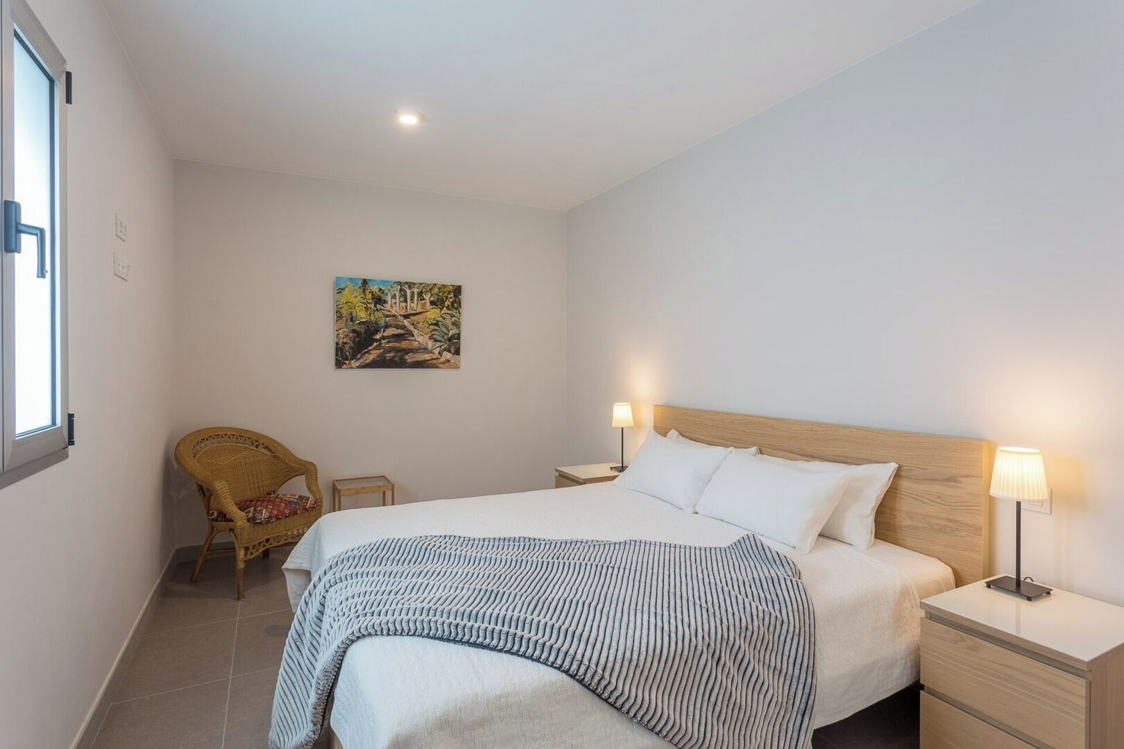 A bedroom with a bed, two bedside tables with lamps, a chair, a painting on the wall, and a window.