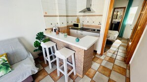 Cocina compacta con una isla central, un fregadero y un microondas. Hay dos taburetes blancos de bar y una planta en maceta.