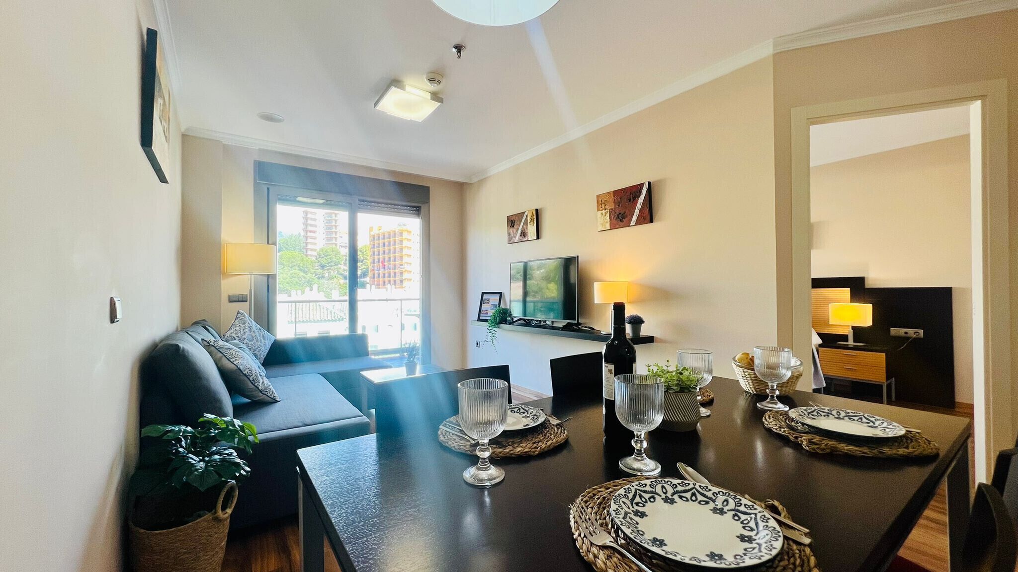 A modern dining area with a dark wooden table set for two, a television on the wall, and a balcony with a view of buildings.