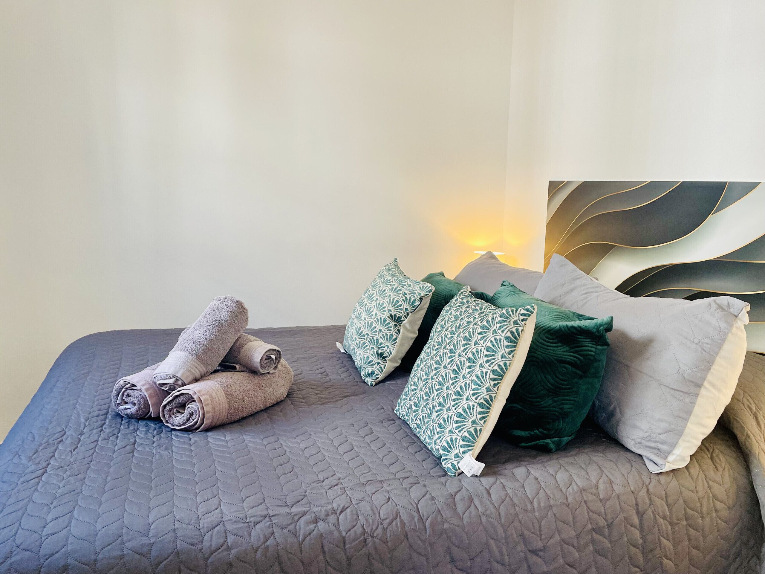 A neatly made bed with rolled towels, green patterned pillows, and a headboard with a unique design.