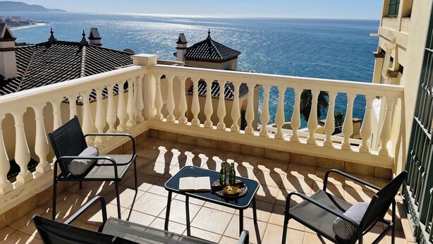 Un balcón con vista al mar, que incluye una mesa con un libro y botellas, y dos sillas.