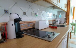 Eine Küche mit Kaffeemaschine, Toaster und Mikrowelle. Ein Spülbecken mit Geschirrgestell zeigt Tomaten. Die Spritzschutzwand weist ein geometrisches Fliesenmuster auf.