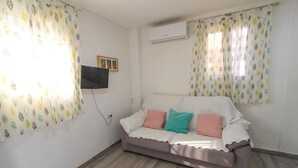 Une petite pièce meublée, comprenant un canapé, un téléviseur à écran plat fixé au mur et un appareil de climatisation.