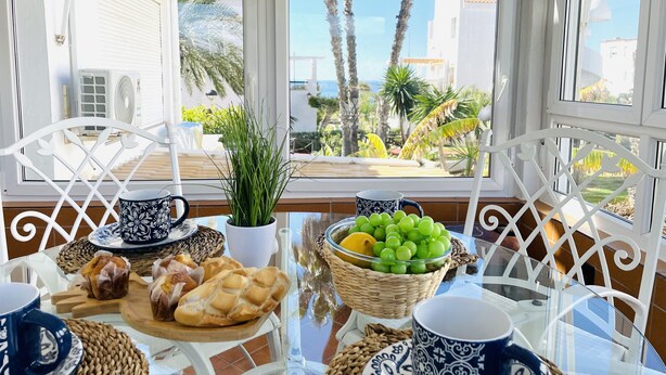 A table set with pastries, a potted plant, and a bowl of fruit, with a view of palm trees and buildings through large windows. - Apartment Urb Laguna Beach Torrox Costa (Torrox Costa)