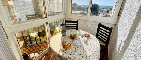 Un comedor con una mesa dispuesta para dos personas, con mantel floral, un plato de cruasanes y un bol de fruta.