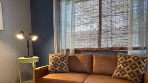Um sofá de couro marrom com almofadas estampadas, uma mesa redonda pequena com uma luminária e uma janela com cortinas leves.