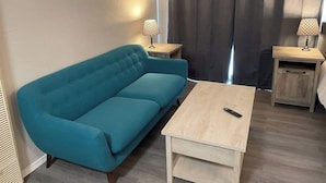 A room with a teal sofa, a wooden coffee table, two bedside tables with lamps, and a window with curtains.