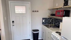 A compact kitchen with a white door, a trash bin, a stove, a sink, and a microwave on a shelf.
