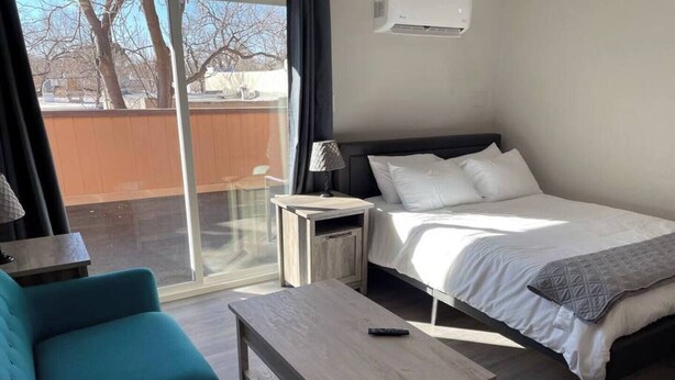 A modern hotel room with a bed, a small table, a sofa, and a window with a view of trees and a fence.