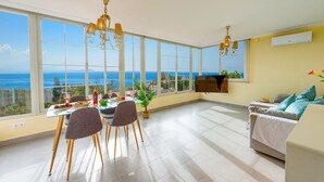 Amplia sala de estar con grandes ventanas que ofrecen vista al mar, una mesa de comedor preparada con comida y un espacio de descanso cómodo con un sofá.