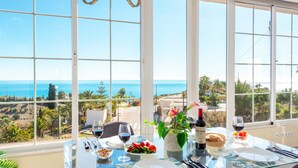 Una mesa de comedor con cubierta de vidrio, copas de vino, una botella de vino y un cuenco de fruta, con vistas a un paisaje costero.