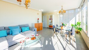 Un salón luminoso con sofá, mesa de centro, mesa de comedor y sillas. Se observan dos candelabros, un aparador y una estantería.