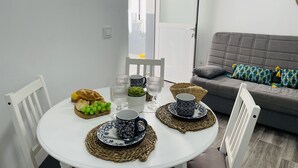 Un comedor con una mesa redonda dispuesta para dos personas, con mantel blanco, dos tazas estampadas, un cuenco de uvas y una cesta de pan.