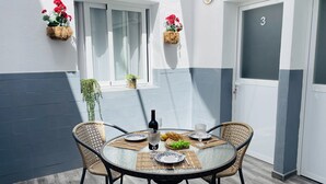 Una mesa redonda con cuatro sillas, una botella de vino y un plato con pan y uvas. Hay dos cestas de flores colgando en la pared, y una puerta con el número 3.
