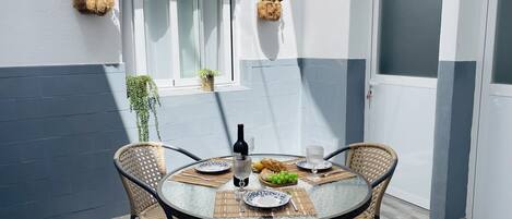 Una mesa redonda con cuatro sillas, una botella de vino y un plato con pan y uvas. Hay dos cestas de flores colgando en la pared, y una puerta con el número 3.
