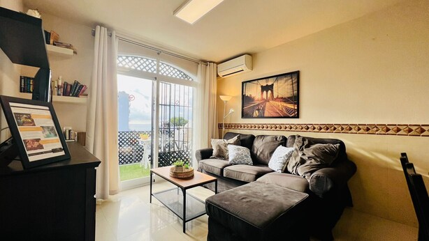 En stue med sofa, skammel og et lille bord med en plante. Der er en balkon med udsigt til en bro og et hvidt gelænder.