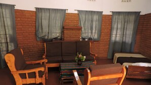 Habitació amb paret de maó, dues cadires de fusta, un sofà, una taula petita amb un vas i un llit.