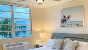 Een slaapkamer met een bed, een plafondventilator, een raam, een wandairconditioner, een bedlampje en een lijst met een afbeelding.