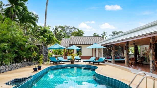 A swimming pool with tiled edges, surrounded by lounge chairs and umbrellas, with a stone wall and tropical vegetation in the background. - Tropical Heavens Garden Samui (Koh Samui)