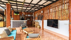 A spacious living area with a stone-tiled floor, wooden furniture, a TV mounted on the wall, and a bamboo wall panel. - Tropical Heavens Garden Samui (Koh Samui)