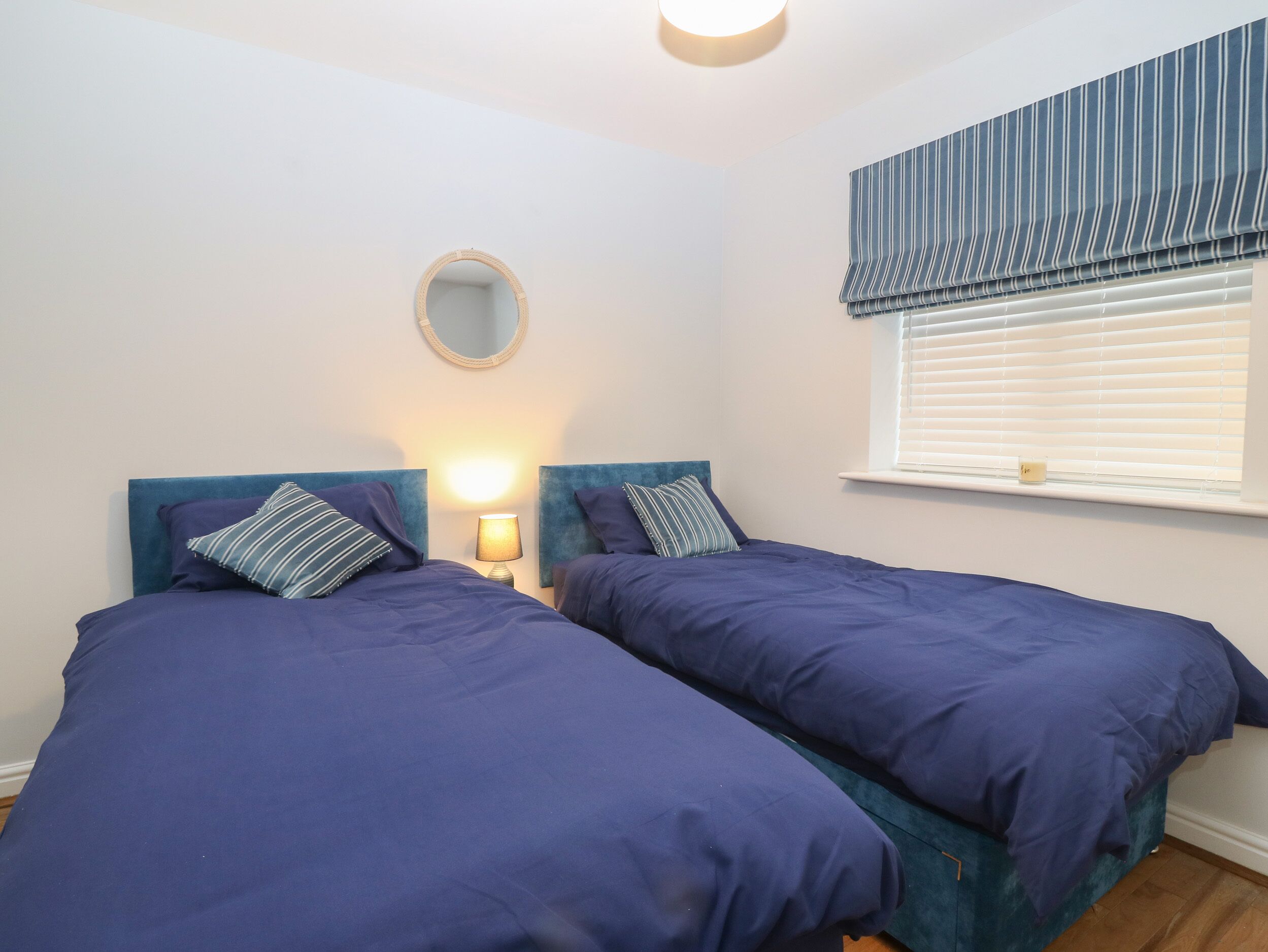 A bedroom with two beds, a round mirror, a bedside lamp, and a window with blinds.