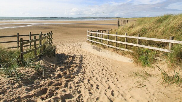 Un sentier de sable qui longe la plage et mène à une clôture en bois d’où l’on a une vue sur l’océan.