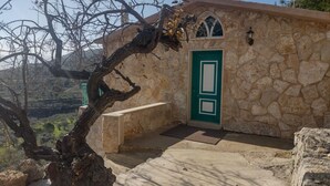 A stone building with a green door and windows, surrounded by trees and a hilly landscape.