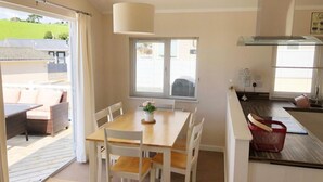 A dining area with a wooden table and white chairs, a window with blinds, and a view of the outside. - Charming 3-bed Chalet in Abersoch (Pwllheli)