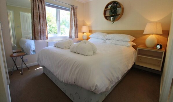 A neatly made bed with white linens and pillows, a bedside table with a lamp and a clock, and a window with curtains.