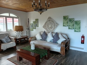 Ein Wohnzimmer mit Holzboden, einer Couch mit gemusterten Kissen, einem Couchtisch, einer Lampe und Bildern an den Wänden.