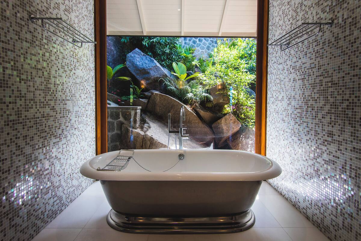 A freestanding bathtub in a bathroom with mosaic tile walls and a view of a garden with rocks and plants.