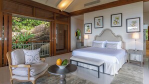 A bedroom with a large bed, a wooden-framed sofa, a small table with a bowl of fruit, and a balcony with a view of greenery. - Villa Salazie (Mahé Island)