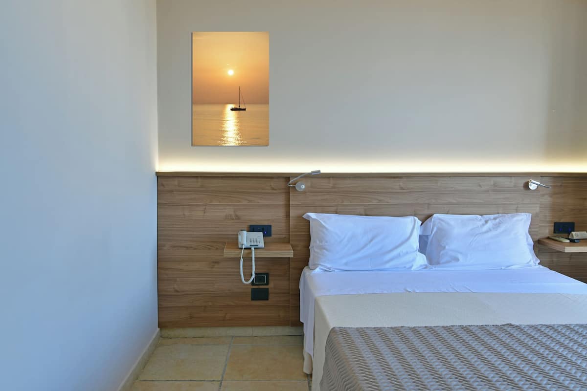 Habitación de hotel con cabecera de madera, cama con ropa blanca, mesita de noche con teléfono y un cuadro colgado en la pared que muestra un atardecer sobre el agua.