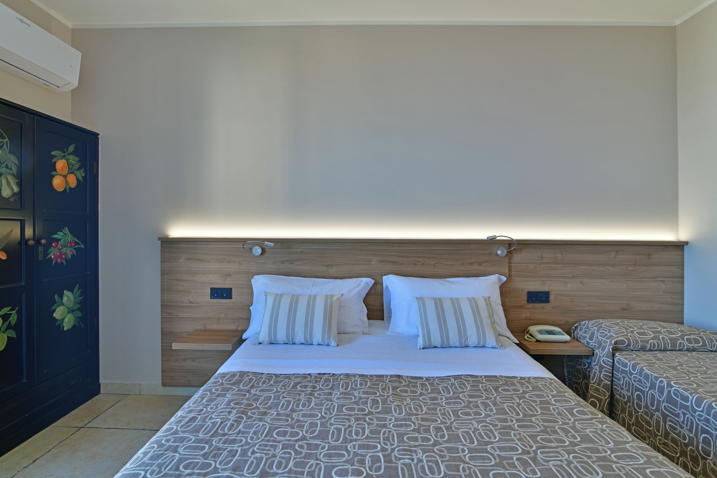 Quarto de hotel com cabeceira de madeira, duas camas com roupa de cama estampada, mesa de cabeceira com telefone e parede com arte decorada com tema de frutas.