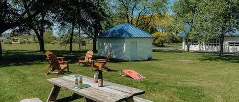 A picnic table with a bottle and a can, surrounded by chairs, in a grassy area with a small shed and trees.