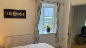 Un dormitorio con cama, un radiador, una ventana con cortinas, un jarrón con flores y una puerta que da a otra habitación.