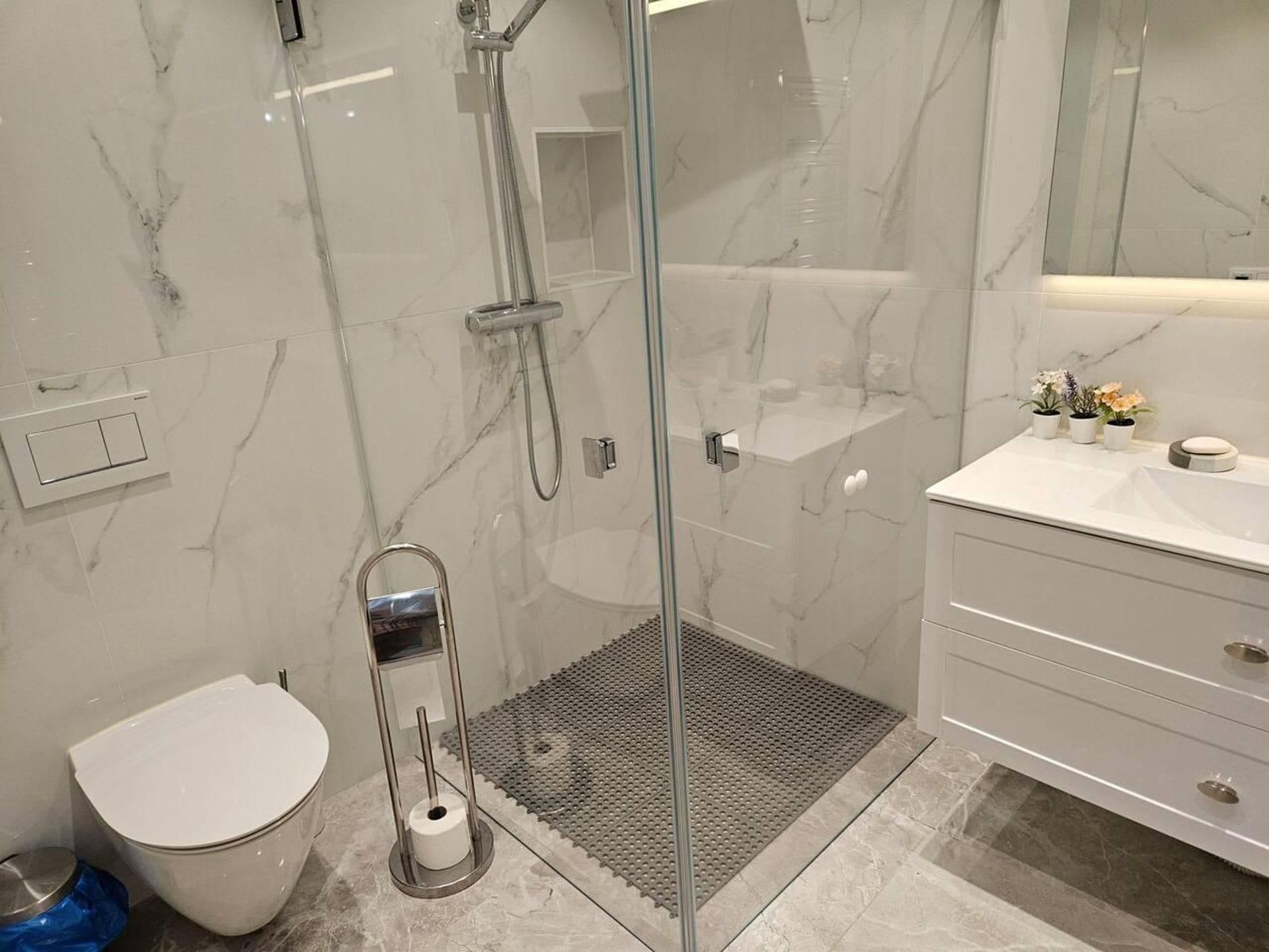 Ett modernt badrum med en dusch som är avskärmad med glasväggar, en toalett, ett handfat med spegel och en liten hylla med toalettartiklar.