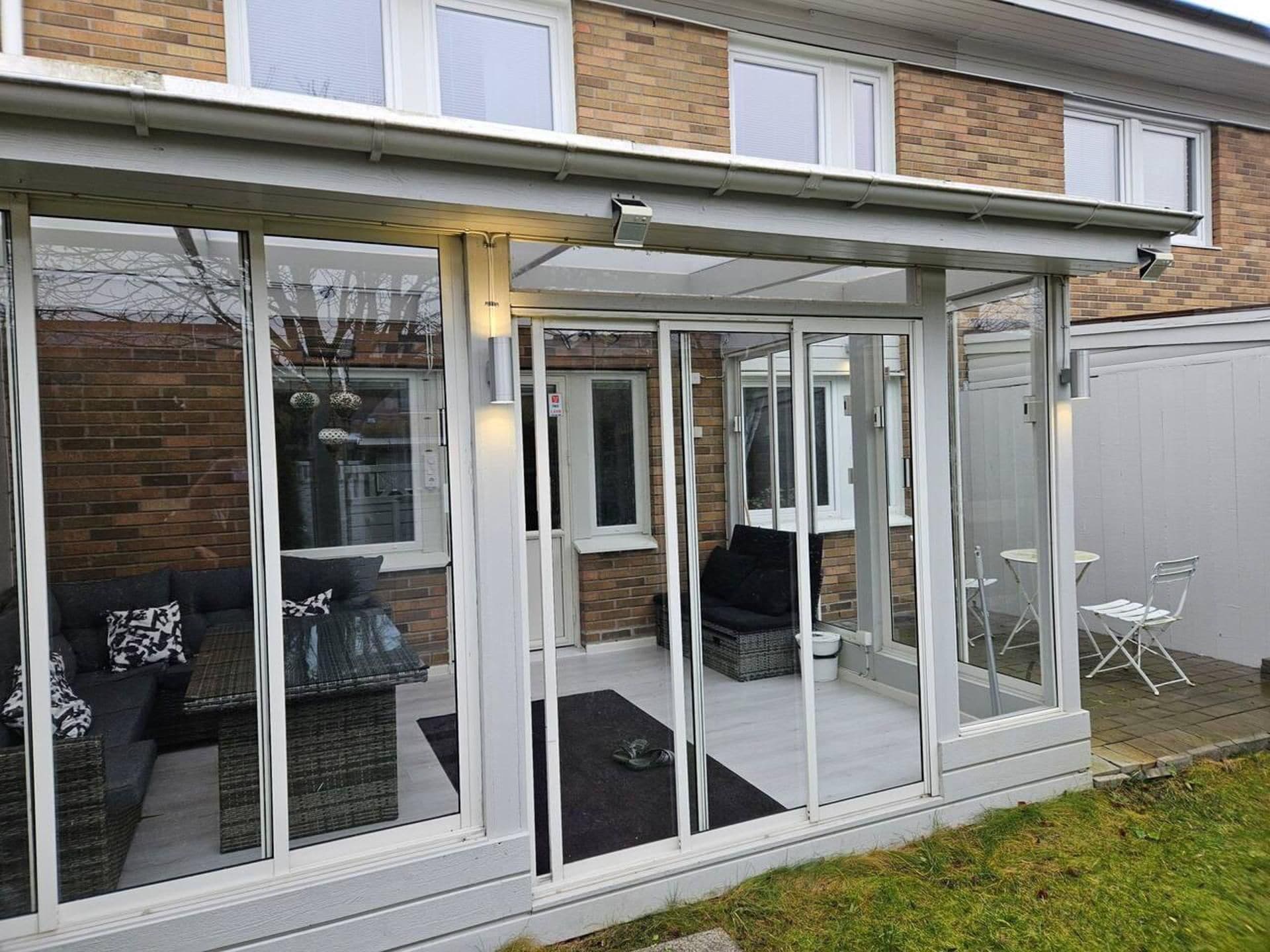 Una casa moderna con un patio cubierto de vidrio que incluye mobiliario exterior y fachada blanca.