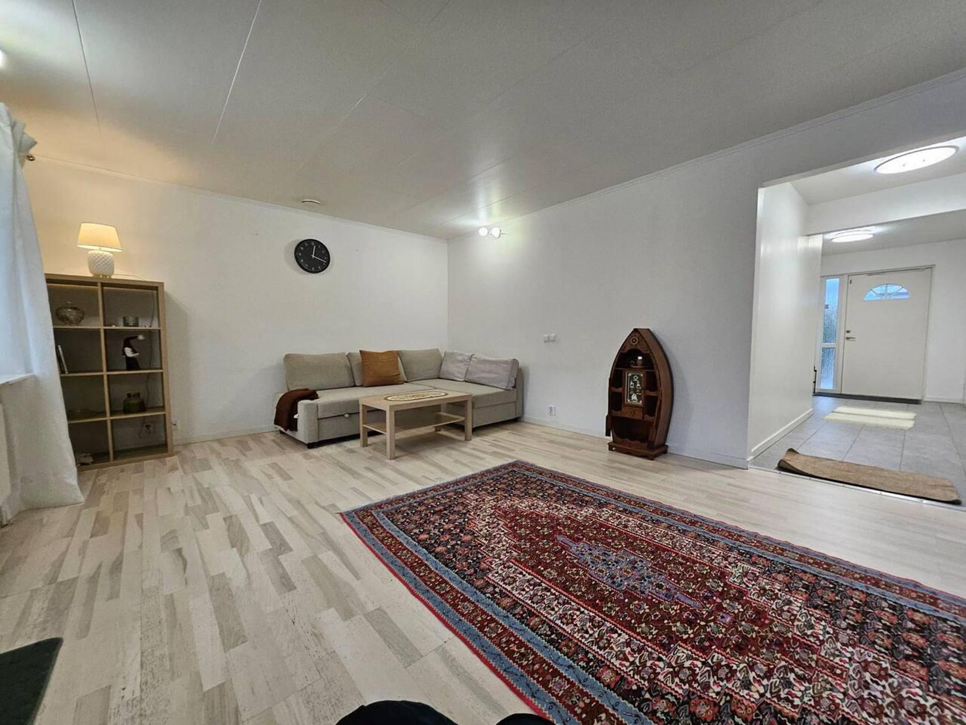 Una sala de estar con una alfombra estampada, un sofá, una mesa de centro, una estantería y un reloj en la pared.