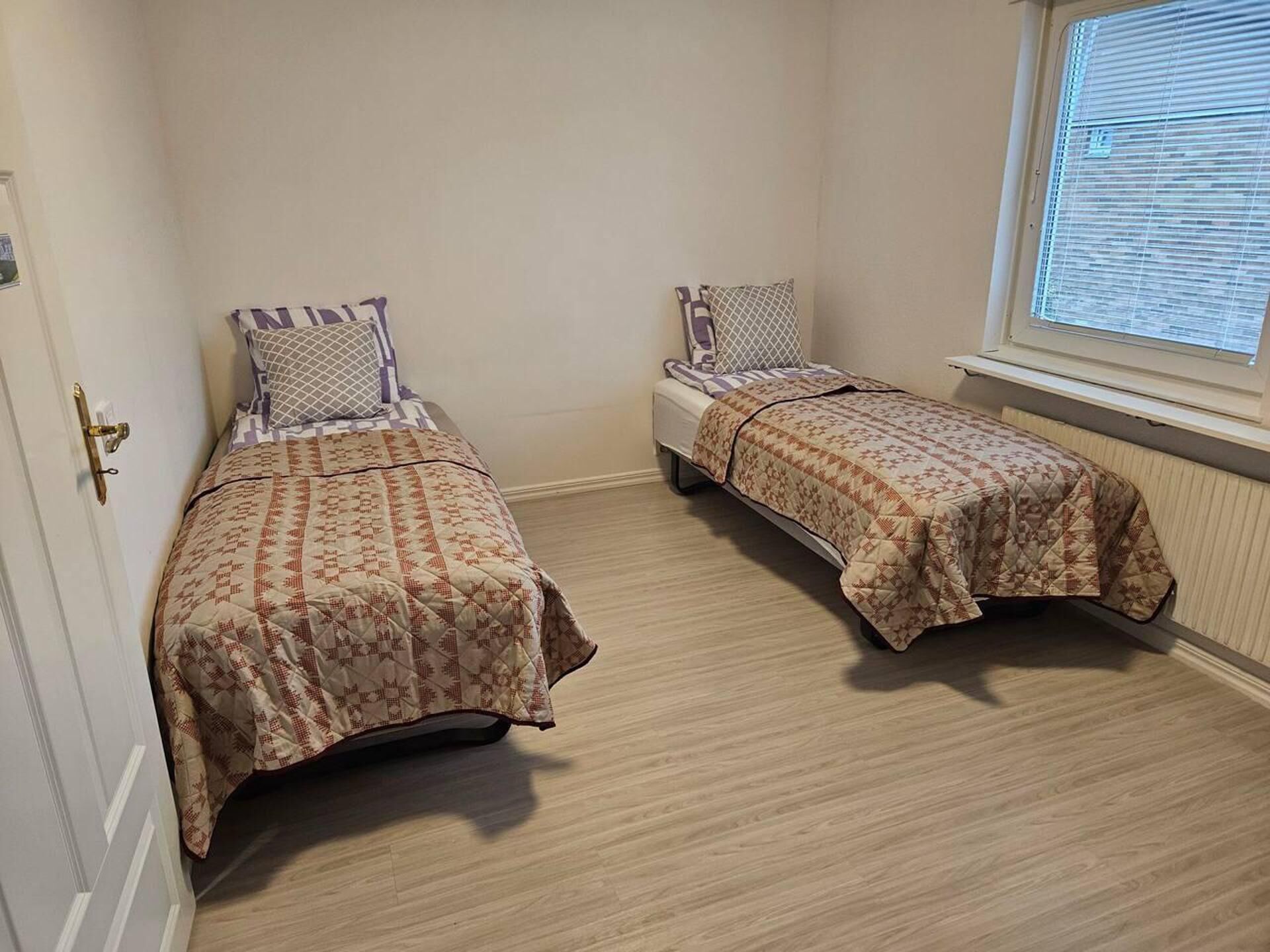 Ein kleines Zimmer mit zwei Einzelbetten, einem Fenster mit Rollläden und einer Tür mit Messingklinke.