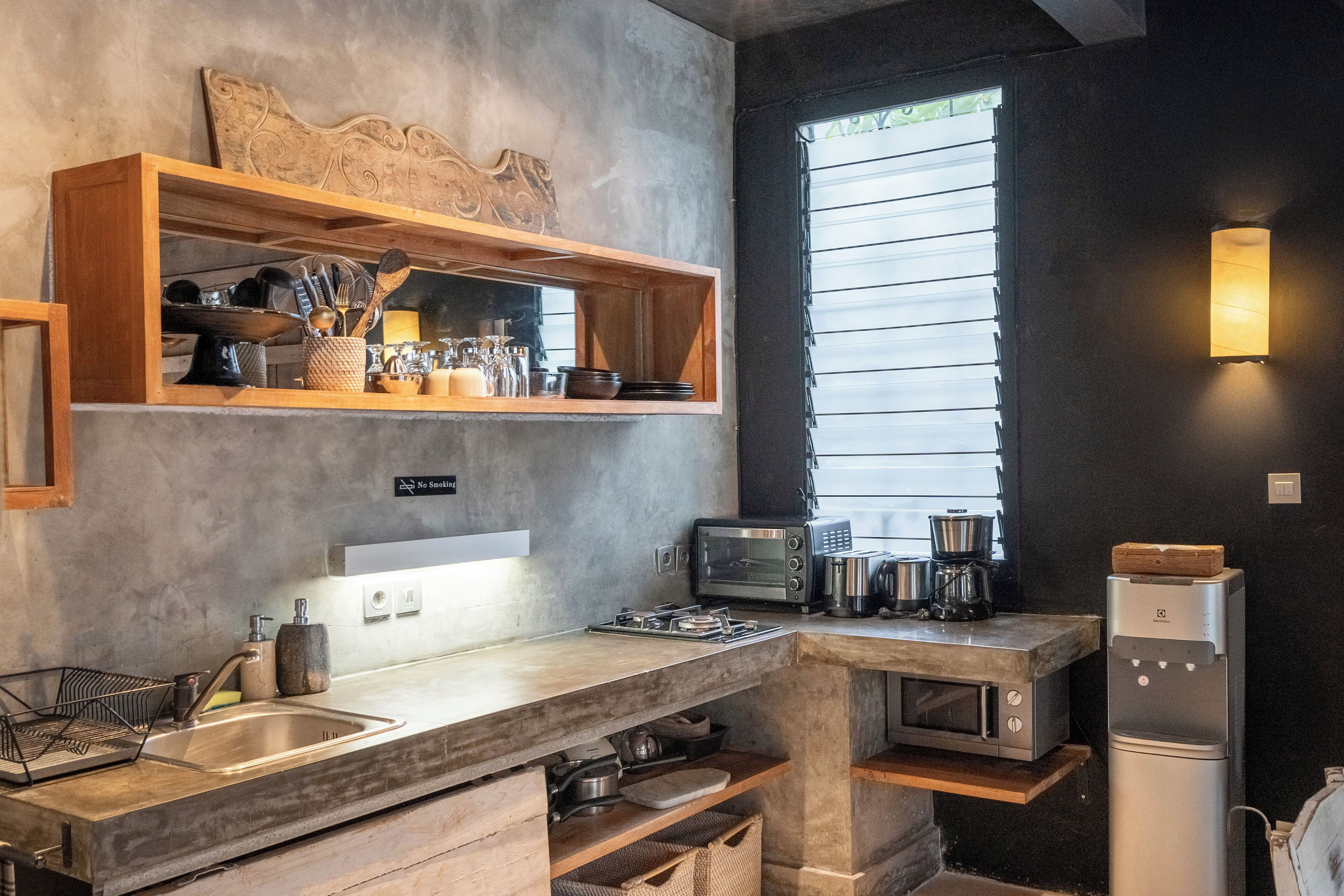 Una cocina moderna con microondas integrado, tostadora y cafetera. Hay un estante de madera sobre el fregadero con utensilios de cocina.