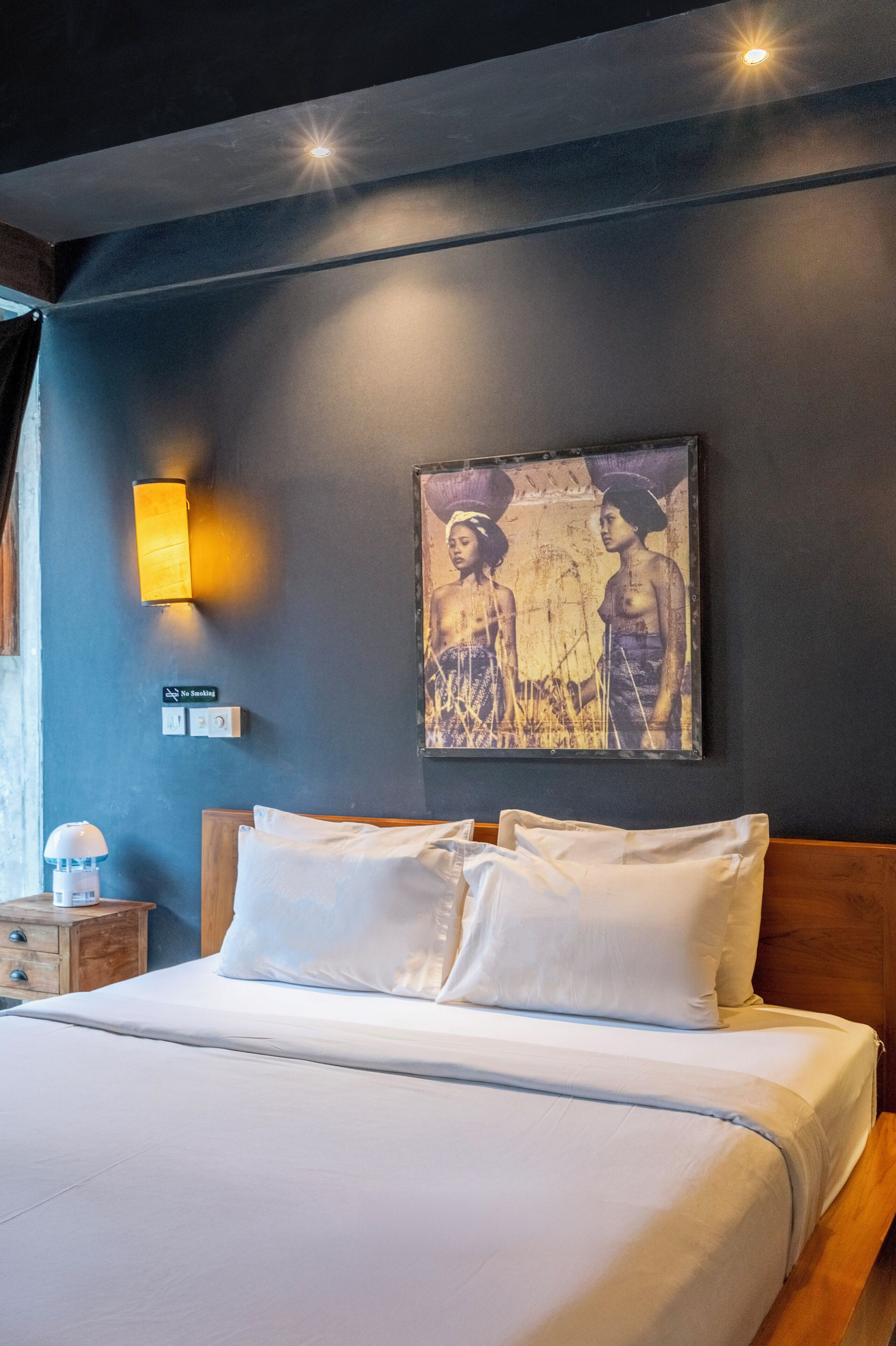 A neatly made bed with white linens, a wooden headboard, a wall-mounted light fixture, and a framed artwork depicting two figures.