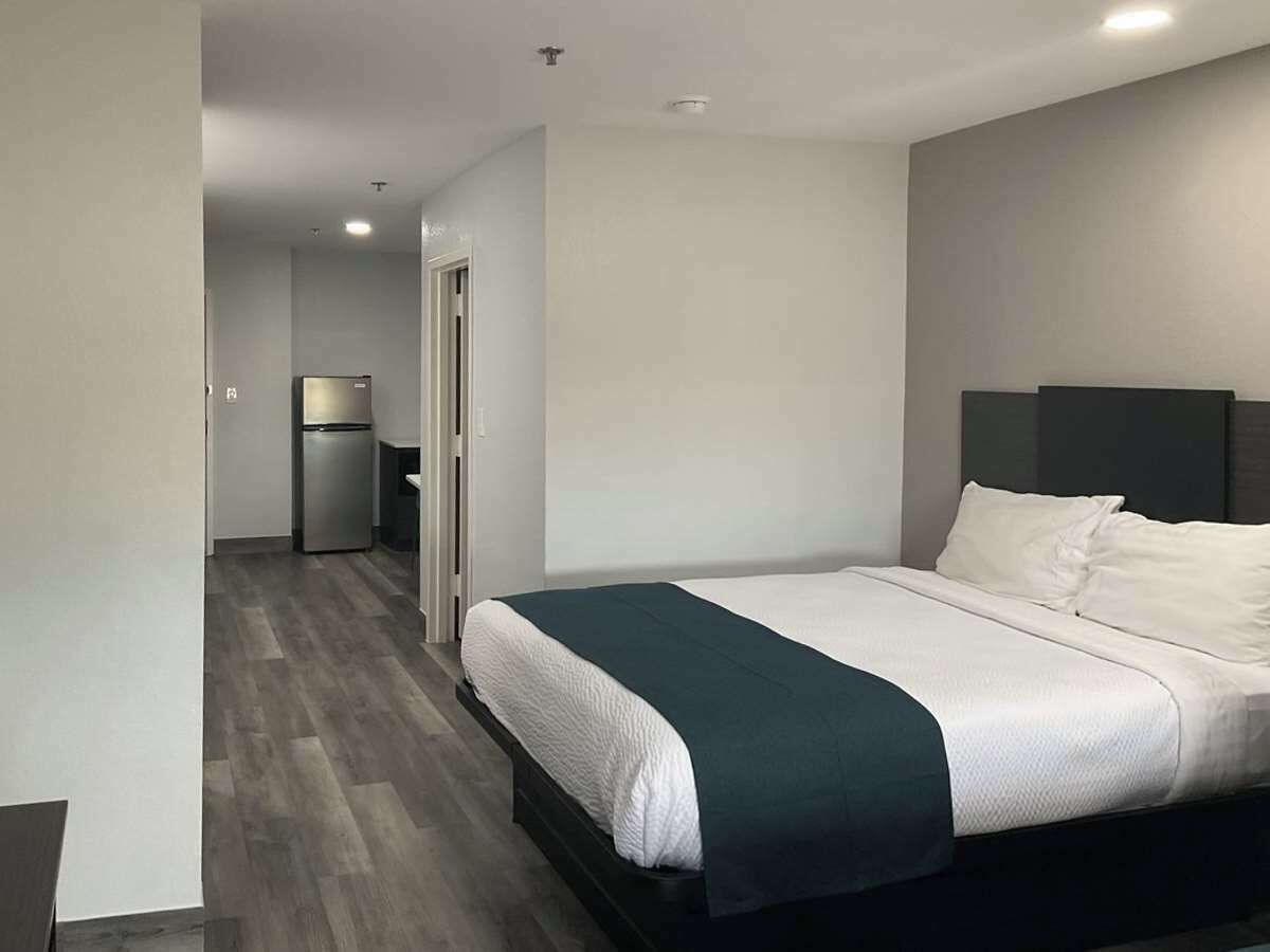 A modern hotel room with a large bed, a dark headboard, and a small kitchenette in the background.