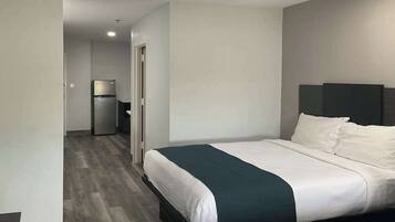 A modern hotel room with a large bed, a dark headboard, and a small kitchenette in the background.