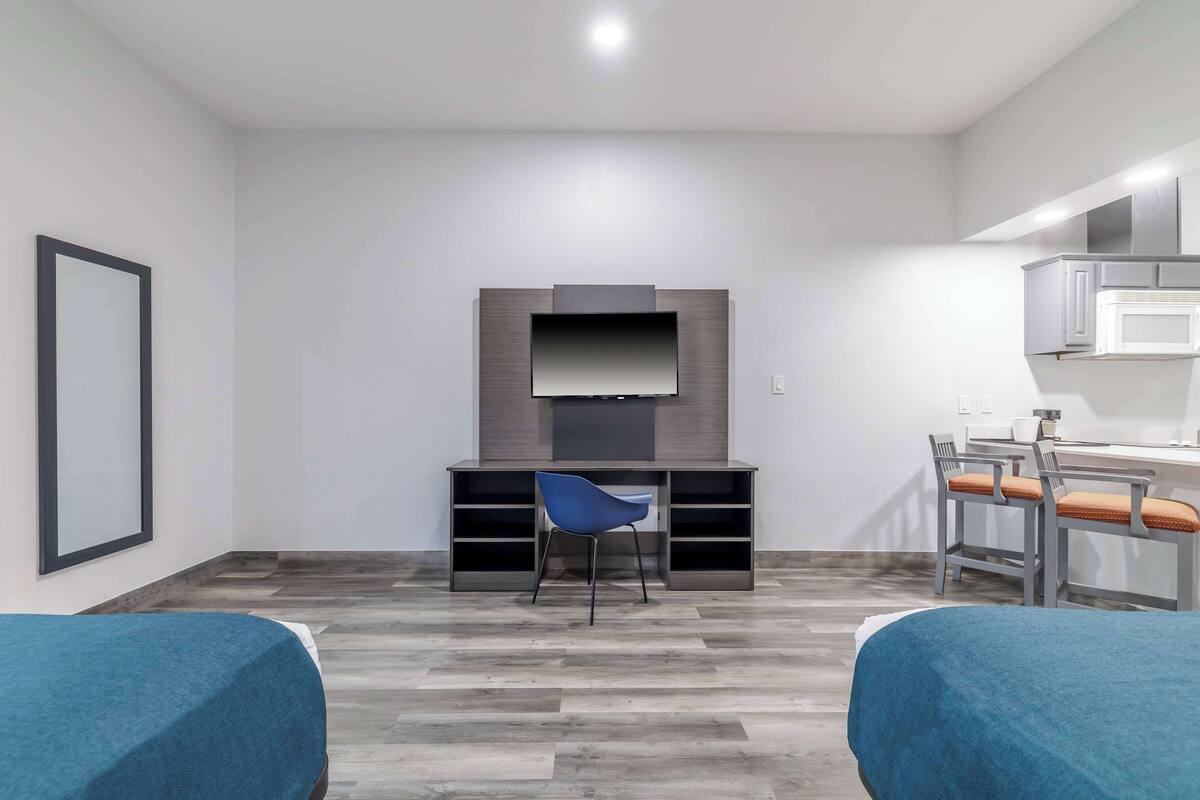 A modern hotel room with a flat-screen TV mounted on a wooden panel, a desk with a chair, and a kitchenette area.