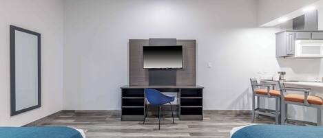 A modern hotel room with a flat-screen TV mounted on a wooden panel, a desk with a chair, and a kitchenette area.