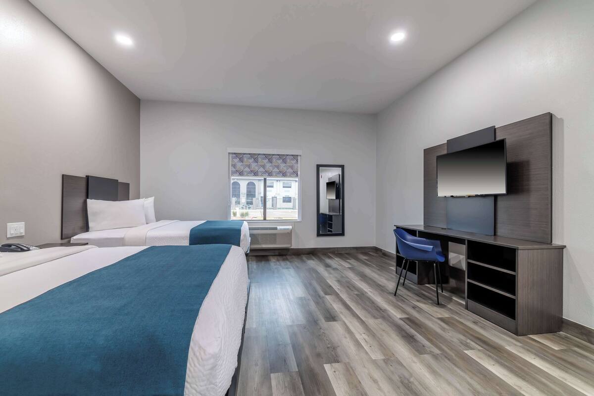 A hotel room with two beds, a desk with a chair, a TV, and a window with a view of buildings.