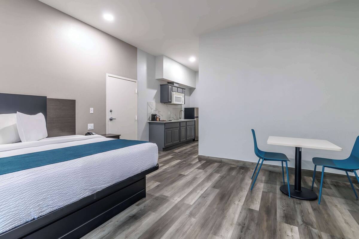 A modern hotel room with a large bed, a small kitchenette, and a dining area with two chairs.